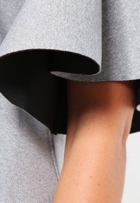 Grått tyg med en slät textur, med en vikt design och kontrasterande svart foder. Fokus på ärmdetaljer och armexponering.