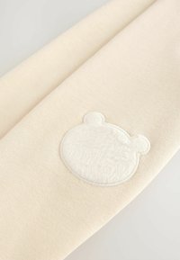 Tessuto color crema con una texture morbida, caratterizzato da un'icona di orsetto ricamata in bianco con orecchie pelose. Design semplice e minimalista.