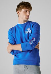 Νεαρός άνδρας με μπλε φούτερ Red Bull Racing, με τα χέρια σταυρωμένα, κοιτάζοντας προς το πλάι μπροστά σε απλή γκρίζα πλάτη.