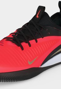 Κόκκινο και μαύρο αθλητικό παπούτσι Nike με μαύρα κορδόνια, χρυσό σήμα Nike, ραβδωτή υφή και μαύρη οδοντωτή σόλα, παρουσιασμένο από κοντινή γωνία.
