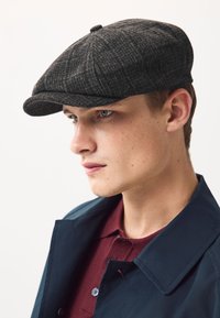 Siva kapa s ribljim kosti uzorkom, zaobljena kruna i kratki, zakrivljeni vrh, nošena od strane mladića s neutralnim izrazom lica.