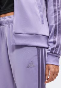 Λιλά αθλητική ζακέτα και παντελόνι Adidas με χαρακτηριστικές τρεις ρίγες και λογότυπο, φορεμένα με μαύρο crop top, δείχνοντας ένα χέρι στο πλάι.