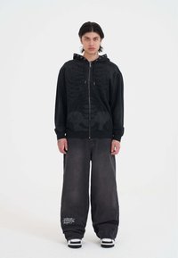 Sudadera negra con estampado de esqueleto, con cremallera en la parte delantera y ojales. Combinada con jeans oscuros holgados que tienen texto gráfico en la pierna izquierda. Zapatillas blancas.