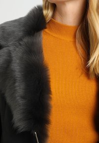Sort jakke med en grå pelskrave over en orange ribbet turtleneck. Fokus på teksturkombinationer og farverne.