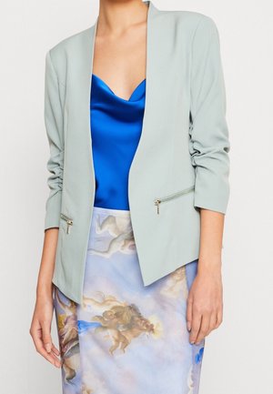 Kvinde iført en lysegrå blazer over en blå satin top og et nederdel med et trykt klassisk cherub- og skydesign.