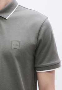 Γκρι πόλο πουκάμισο με υφή υφάσματος, λευκές λεπτομέρειες στο γιακά και τετράγωνο λογότυπο στο κέντρο του στήθους.