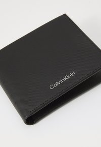 Sort bifold tegnebog lavet af struktureret syntetisk materiale, med en sølv "Calvin Klein" logo nederst til højre.