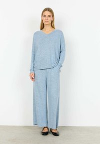 Femeie în picioare purtând un pulover tricotat moale albastru deschis cu decolteu în V și pantaloni largi asortați, cu pantofi negri Mary Jane.