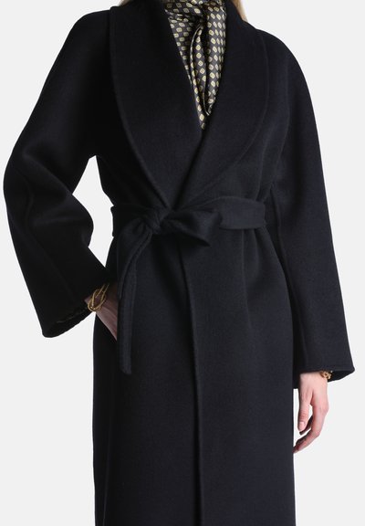 Manteau en laine noire ceinturé par-dessus une écharpe en soie à motifs, porté par une personne avec la main dans la poche et des bracelets torsadés en corde dorée.