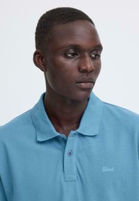 Polo bleu clair en tissu texturé, avec un col classique, deux boutons et un discret logo brodé sur la poitrine.