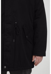 Svart parka-jakke med glidelås foran, snøring i midjen og en sidelomme med trykknapp. Glatt tekstur, avslappet passform.