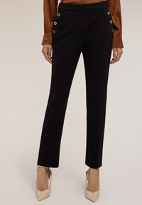 Pantalons noirs ajustés avec poches latérales, dotés de grandes œillets dorés, au design droit et à la texture lisse, associés à des talons clairs.