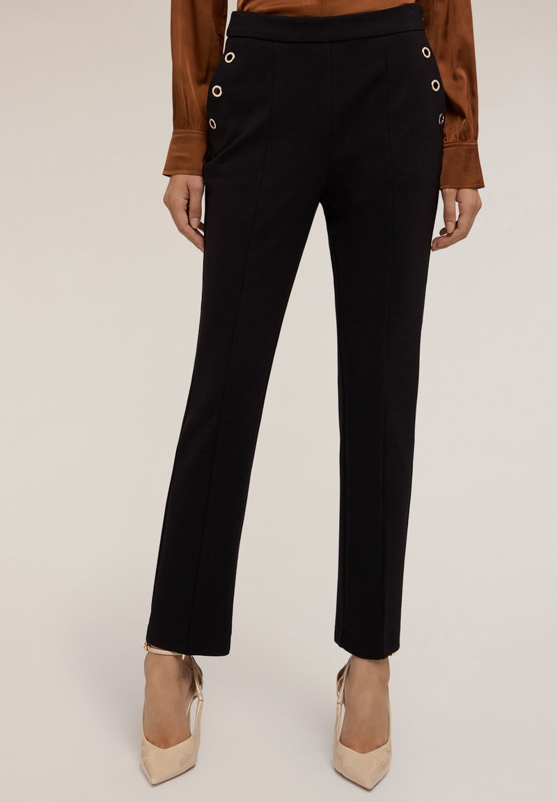 Pantalons noirs ajustés avec poches latérales, dotés de grandes œillets dorés, au design droit et à la texture lisse, associés à des talons clairs.