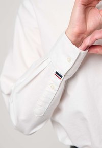 Person wearing a white buttoned shirt adjusting the cuff with a small red, white, and blue striped tag near the wrist.