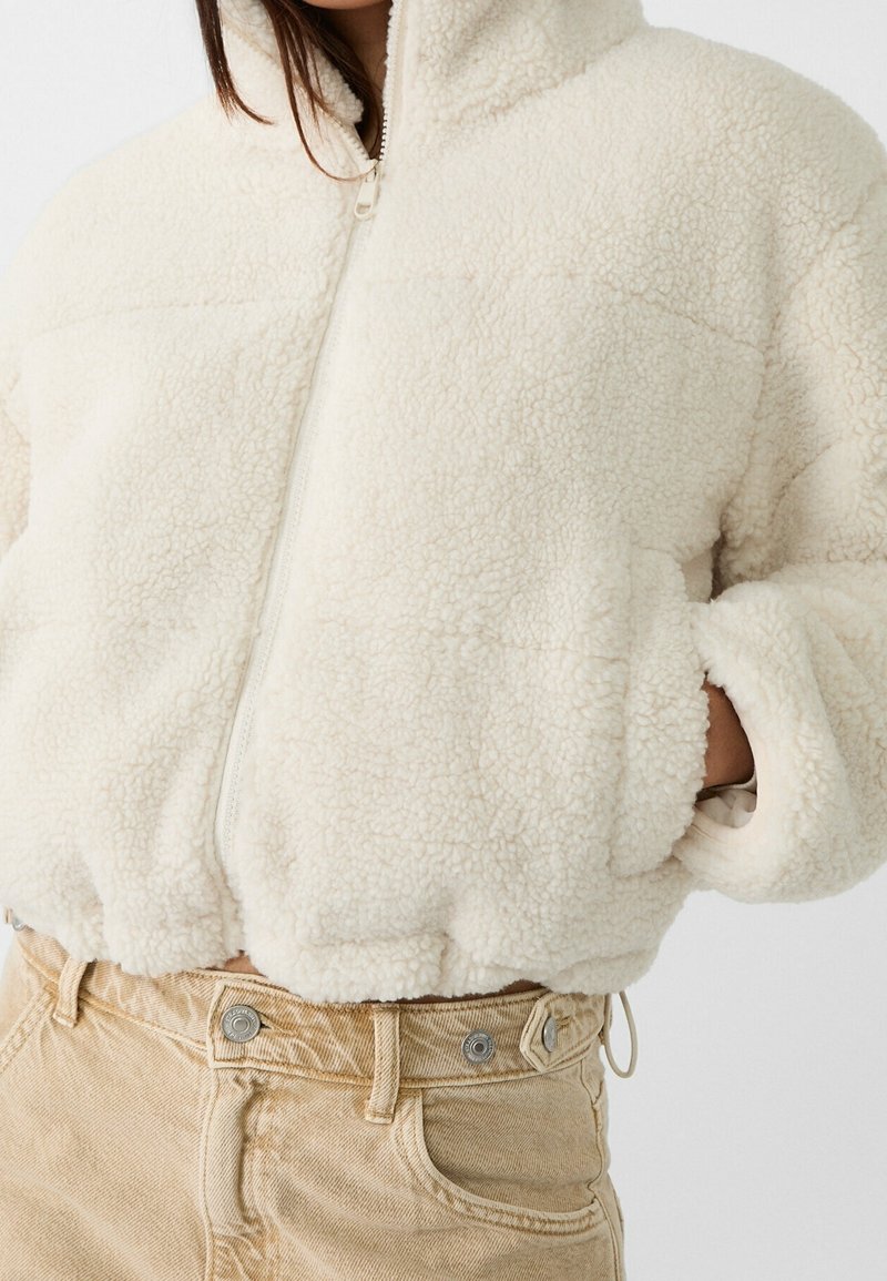 Femme portant une veste sherpa crème zippée avec les mains dans les poches et un pantalon beige taille haute.