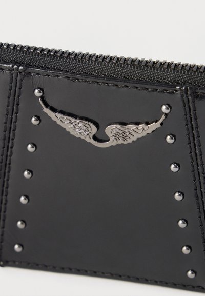 Τμήμα πορτοφολιού από μαύρο δέρμα με μεταλλικό έμβλημα φτερού και μικρά ασημένια τρουκς, κοντινή λήψη που δείχνει ραμμένες άκρες και φερμουάρ.