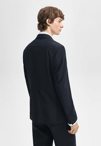 Marineblauwe colbert met een enkele split, notch-revers en knopen met drie knoopsgaten. Gladde stof en getailleerde pasvorm, met minimaal schouderpadding.