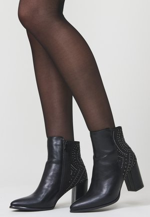 Piernas en medias negras transparentes que llevan botas de cuero negras hasta el tobillo con detalles de tachuelas y tacones cuadrados sobre un fondo liso.