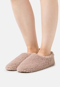 Fuzzy, light brown slippers with a low cut design, soft texture, and pointed toe shape. Positioned on bare feet with a smooth finish.