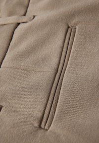 Beżowa tkanina o teksturowanym wykończeniu, z pojedynczą pionową kieszenią po lewej stronie oraz czystym, prostym wzorem.
