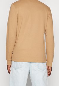 Maglietta a maniche lunghe a coste color marrone chiaro, con scollatura tonda e maniche a polsino. Abbinata a jeans blu chiaro.