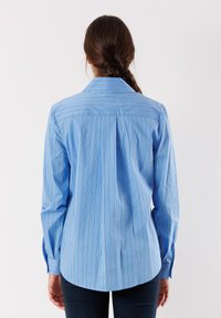 Camicia blu a maniche lunghe con righe verticali bianche, dotata di un colletto classico, polsini abbottonati e orlo arrotondato. Tessuto di cotone liscio.