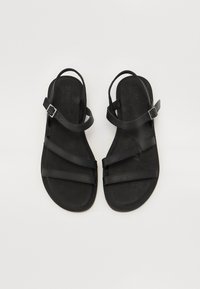 Sandalias planas negras con múltiples correas y una hebilla. Fabricadas con material suave y textura lisa. Diseño minimalista, con los dedos y el talón al descubierto.