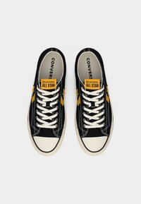 Μαύρα πάνινα παπούτσια με κίτρινα αστέρια, λευκά κορδόνια και ετικέτες λογότυπου. Διαθέτουν λαστιχένιο κάλυμμα μύτης και ραμμένες λεπτομέρειες.