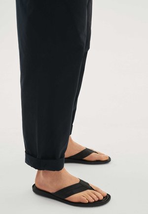 Person trägt schwarze kurze Hosen und schwarze Flip-Flop-Sandalen und steht auf einer hellen Oberfläche.