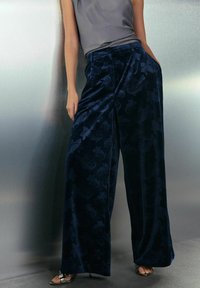 Woman wearing dark blue velvet wide-leg pants with floral pattern, gray sleeveless top, and silver open-toe heels, standing against metallic wall.