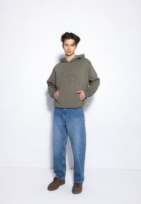 Zielono-oliwkowy hoodie z wypukłym napisem "LOKALNI BOHATEROWIE", w połączeniu z luźnymi niebieskimi dżinsami i brązowymi butami. Gładka tekstura, casualowy design.