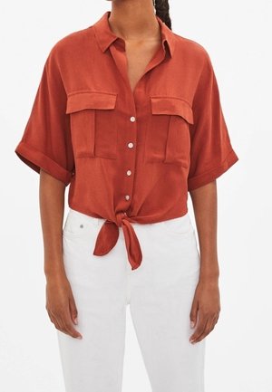 Košile s krátkým rukávem v rezavě oranžové barvě, zapínaná na knoflíky s kapsami na hrudi, uvázaná v pase, nošená s bílými kalhotami s vysokým pasem.