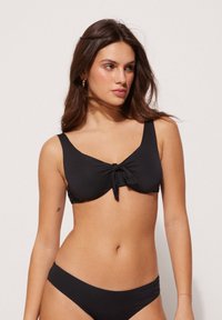 Schwarzer Bikini-Oberteil mit einem Knoten-Detail in der Mitte, breiten Trägern und einer glatten Textur. Kombiniert mit passenden Bikini-Unterteilen.
