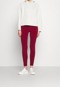 Jasno beżowa bluza z kapturem z przednią kieszenią, połączona z czerwonymi legginsami i białymi trampkami, o dopasowanym kroju i gładkiej fakturze.