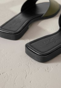 Sandales plates noires à finition lisse, avec une bride accent verte, forme carrée de l'orteil, et une semelle texturée sur une surface claire.