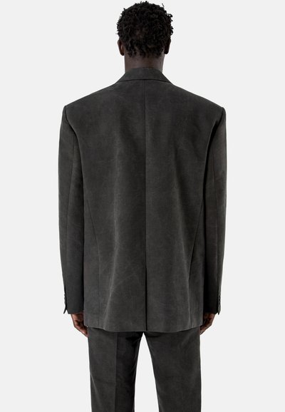 Mężczyzna stojący tyłem, ubrany w ciemnoszary, oversize'owy blezer oraz dopasowane spodnie na jasnym, jednolitym tle.