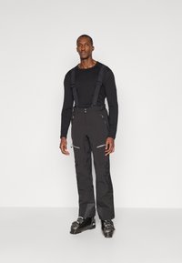 Schwarze Skihosen mit verstellbaren Trägern, Reißverschlusstaschen und einer glatten Textur. Getragen mit schwarzen Skischuhen und einem enganliegenden Langarmshirt.