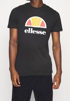 Mand iført sort kortærmet t-shirt med gul og orange halvcirkellogo og teksten "ellesse" på fronten, kombineret med mørke bukser.