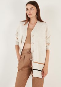 Light beige cardigan with a soft texture, featuring a v-neck, button closure, and horizontal stripes in brown and black at the hem.