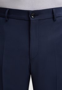 Nahaufnahme von dunkelblauen maßgeschneiderten Hosen mit befestigtem Bund und Gürtelschlaufen, getragen mit einem weißen Oberteil.