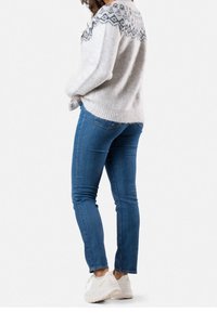 Femme portant un pull gris à motif, un jean bleu et des baskets blanches, debout les mains dans les poches, légèrement de profil.