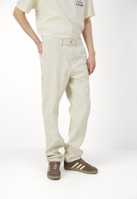 Pantaloni cargo bej din bumbac, cu o croială relaxată, având buzunare laterale, închidere cu nasture și detalii vizibile ale cusăturilor.