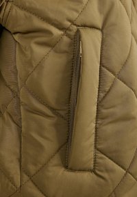 Tissu matelassé vert olive avec une poche verticale zippée partiellement ouverte, montrant les dents de la fermeture éclair et la tirette.