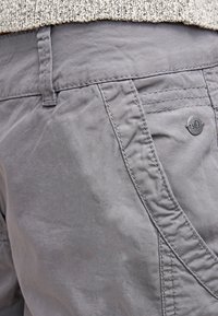 Pantalones grises con un corte relajado, una textura sutil y un pequeño logo metálico redondo en el bolsillo trasero. Detalles de costura a lo largo de las costuras.