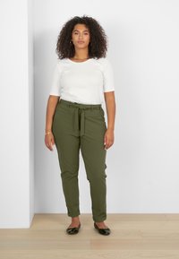 T-shirt blanc à manches courtes et pantalon ajusté vert olive avec une ceinture. Le modèle se tient sur un sol en bois clair devant un fond blanc.