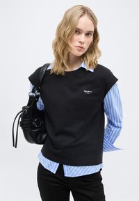 Νέα γυναίκα με κυματιστά ξανθά μαλλιά φορά μαύρο αμάνικο τοπ πάνω από μπλε ριγέ πουκάμισο, κρατώντας μαύρη δερμάτινη τσάντα ώμου σε λευκό φόντο.