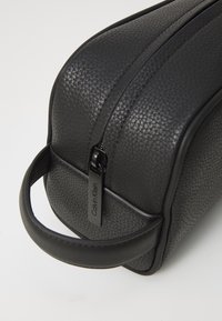 Trousse de maquillage noire en cuir texturé. Présente une forme arrondie, une fermeture éclair sur le dessus et une petite poignée pour un transport facile.