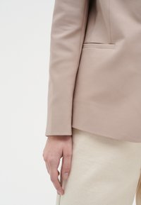 Ljus beige blazer med en slät yta, skräddarsydd passform och en enda ficka. Bärs över ljusfärgad tyg, vilket visar rena linjer.