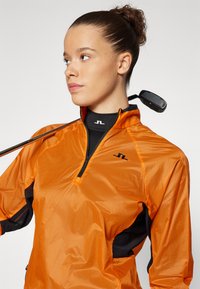 Oranžna jakna na zadrgo s črnimi poudarki, izdelana iz lahkega, teksturiranega materiala. Ima logo na prsih in ovratnik. V ozadju je golf igrišče.