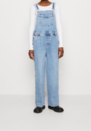 Lichtblauwe spijkerbroek-overalls met verstelbare bandjes, voorzien van een zak aan de voorkant en knoopaccenten aan de zijkant. Draagt over een wit shirt met lange mouwen.